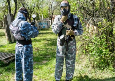 Dvije osobe u maskirnim odijelima i zaštitnim maskama na paintball terenu drže paintball puške, okruženi zelenilom i drvenim preprekama.