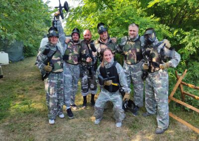 Grupa ljudi u maskirnim uniformama i zaštitnoj opremi drži paintball puške i pozira na otvorenom prostoru s drvećem u pozadini.