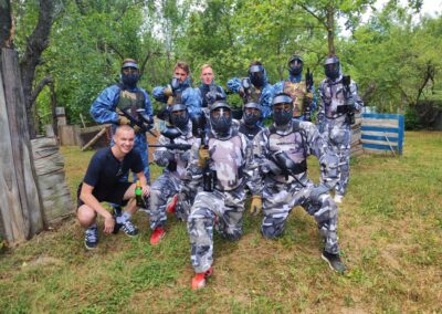 Grupa ljudi u zaštitnoj maskirnoj opremi pozira s opremom za paintball na otvorenom, okružena drvećem i preprekama, dok jedan mladić u crnoj majici kleči ispred njih s bocom pića u ruci.