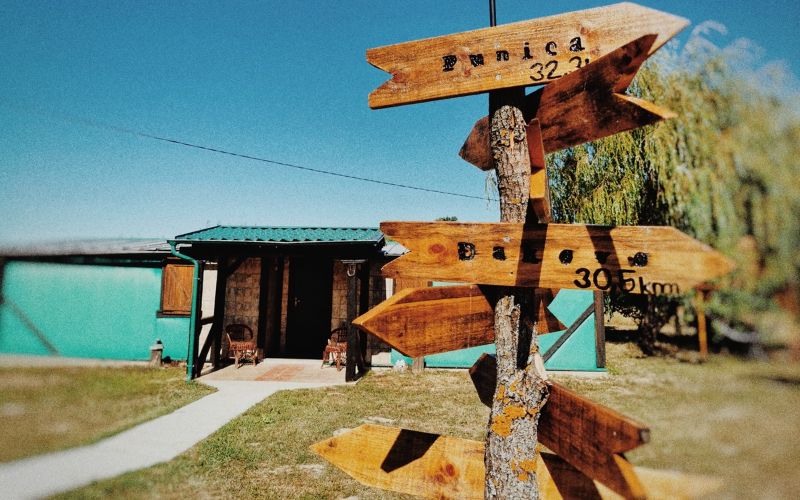 Drvena strelica na stablu pokazuje udaljenost do obližnjih gradova, ispred skromne kuće za odmor s terasom i zelenim krovom, okružena travnjakom i stablima pod vedrim nebom.